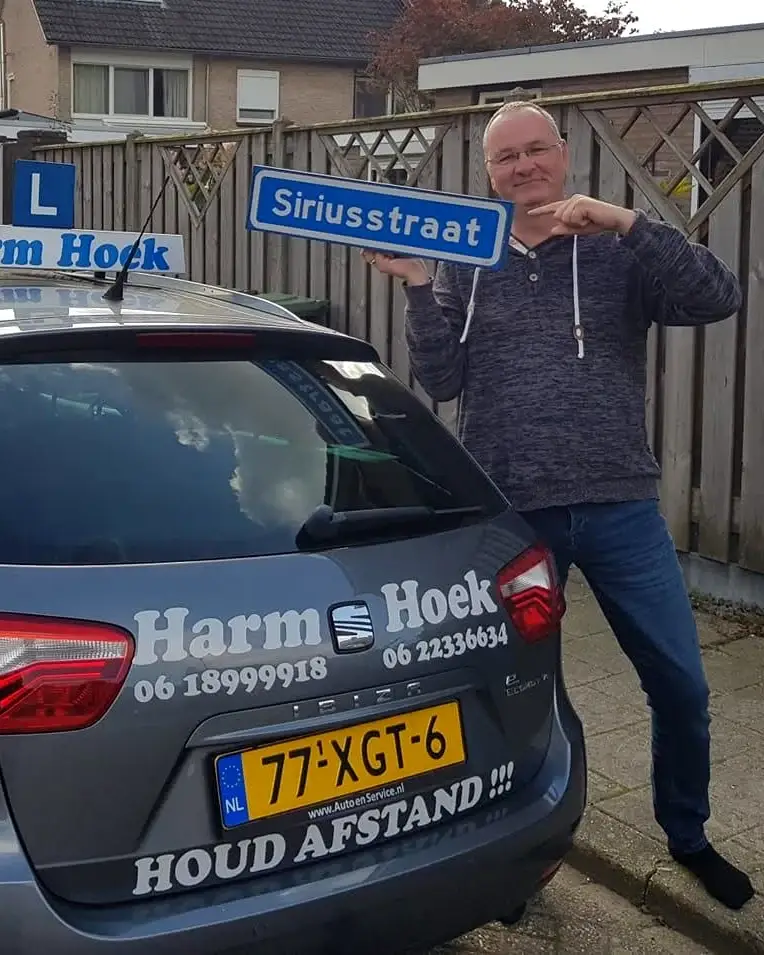 Rijinstructeur Harm Hoek bij de lesauto, met het straatnaambord van de Siriusstraat